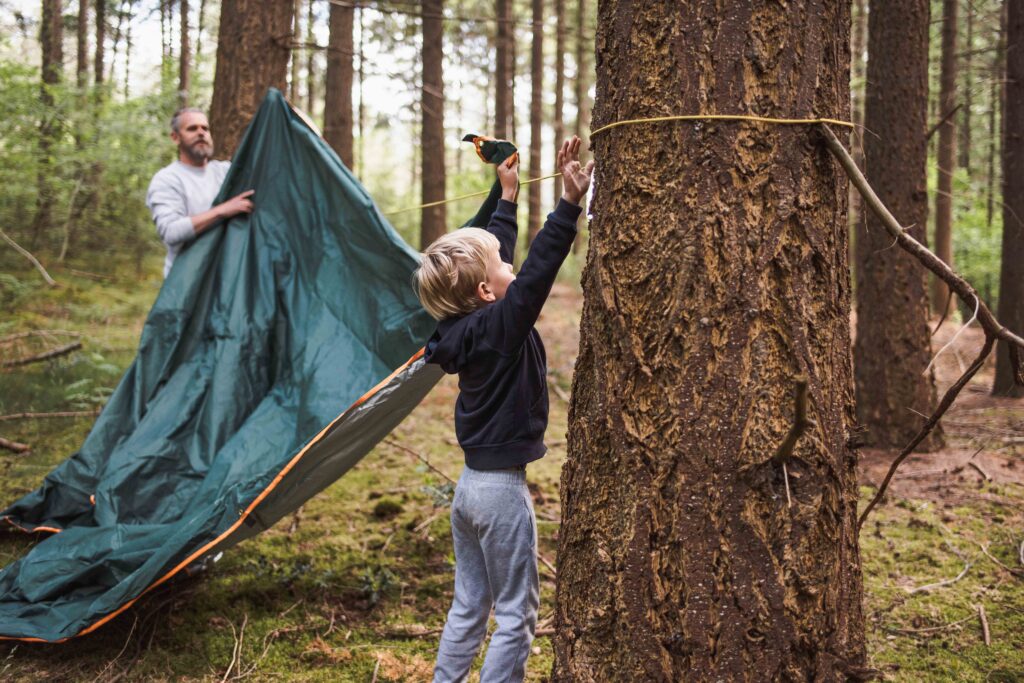 Een kind knoopt een touw om een boom om een zeil op te hangen, terwijl een volwassene het zeil vasthoudt in een bos. Beiden zijn gefocust op hutten bouwen, omringd door bomen en groen, en omarmen de vreugde van ouderlijke momenten tijdens hun buitenavontuur.