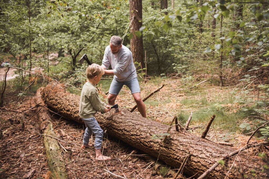 Een man en een kind zijn bezig met ouderlijk houthakken in een bos, waarbij ze een omgevallen boom omhakken. De man, gekleed in een grijs shirt en korte broek, hanteert een bijl terwijl het kind, gekleed in een groen jasje en jeans, een andere bijl hanteert. Ze worden omgeven door weelderig groen.