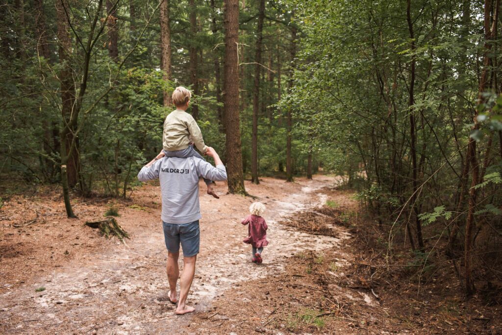 Een persoon loopt op blote voeten over een bospad, met een kind op zijn schouders, wat de geest van ouder-kind trainingen belichaamt. Een ander kind, in een roze jurk, verkent de omgeving. Het pad wordt omarmd door bomen en groen, wat een serene omgeving creëert die perfect is voor buitenspelen en wilde groei.