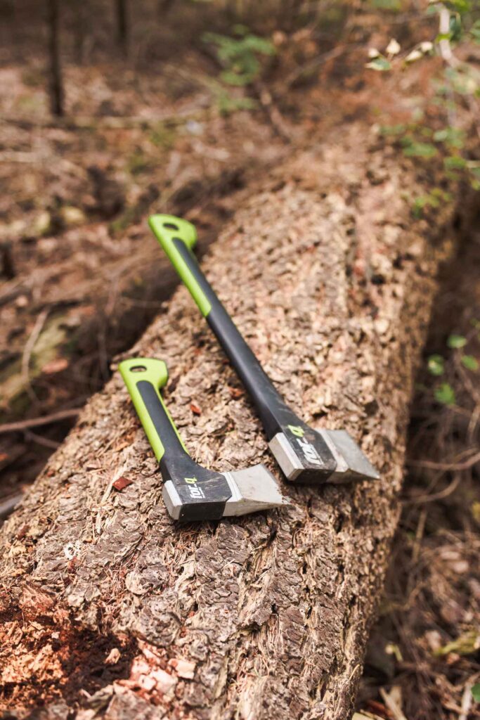 Twee bijlen met groene en zwarte handvatten rusten op een grote, omgevallen boomstam in het bos. Perfect voor hutten bouwen avonturen voor oudere kinderen, de achtergrond is rijk aan droge bladeren en wazige bostinten.