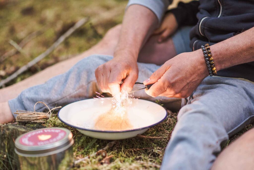 Een persoon gebruikt een vuurstarter om vonken te maken boven een kom op het met mos bedekte gras, wat doet denken aan buiten spelen adventures. Ze zitten met gekruiste benen in casual kleding, met een klein blikken bakje in de buurt. De arm van een ander persoon is gedeeltelijk zichtbaar op de achtergrond, wat duidt op collaboratief hutten bouwen.