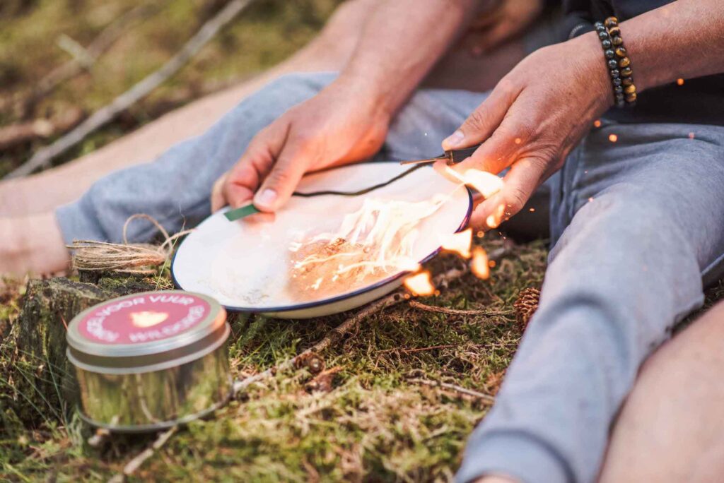 In het bos steekt een persoon die *buitenspelen* doet een vuur aan op een metalen plaat met een vuurstarter en vuursteen. Zittend op de grond, met een armband om, worden ze omringd door de rust van de natuur. In de buurt draagt een blik met het label "Nordic Vulp" bij aan hun avontuurlijke geest.