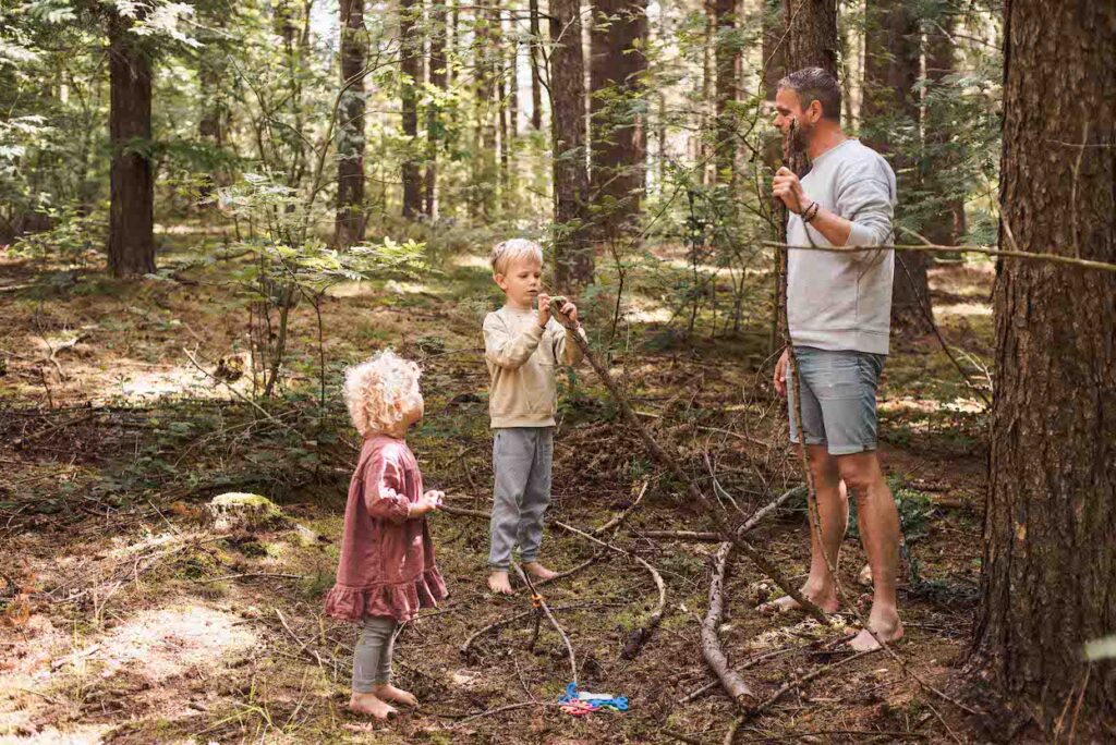 Een man en twee kinderen verzamelen takken in een bos. De man en het oudere kind zijn druk bezig hutten bouwen, stokken aan elkaar vastbinden, terwijl het jongere kind, in een roze jurk, toekijkt. Verwikkeld in ouder-kind binding, zijn ze op blote voeten te midden van het weelderige groen.