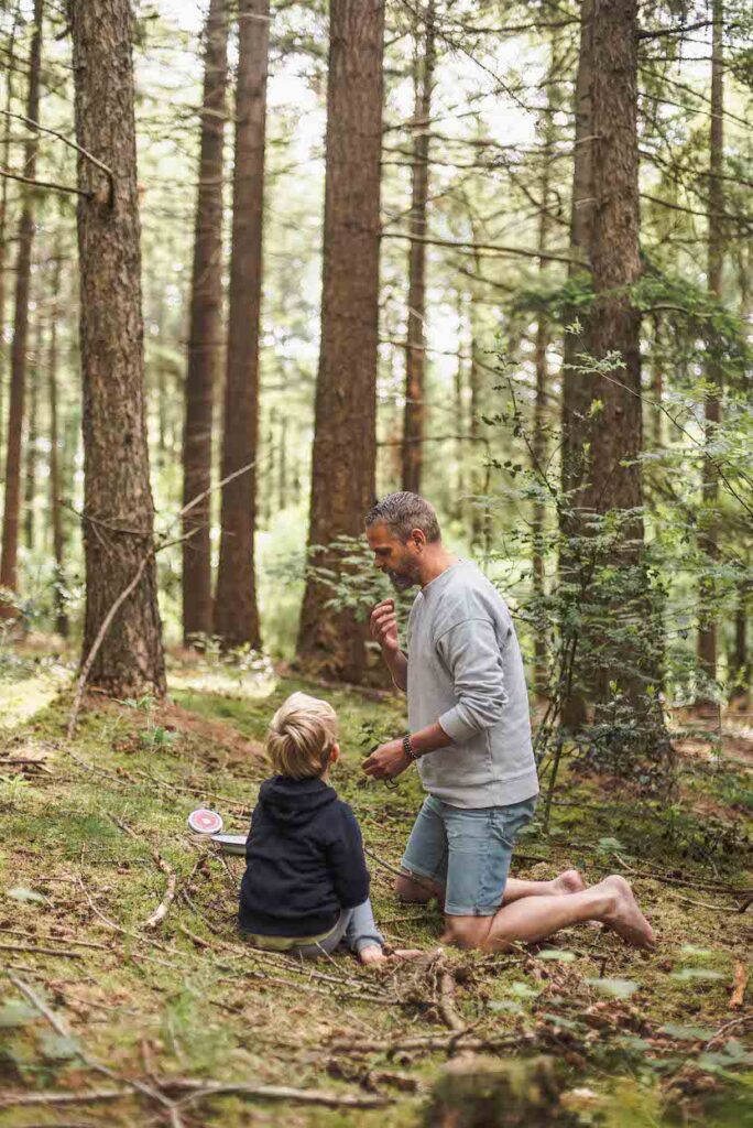 Een man en een kind knielen in een bos, omringd door hoge bomen en groen gebladerte. Ze doen aan ouder-kind trainingen en genieten van een picknick, waarbij de man een voorwerp vasthoudt en het kind aandachtig toekijkt op een kleedje. Het is een zonnige dag, perfect voor buitenspelen.