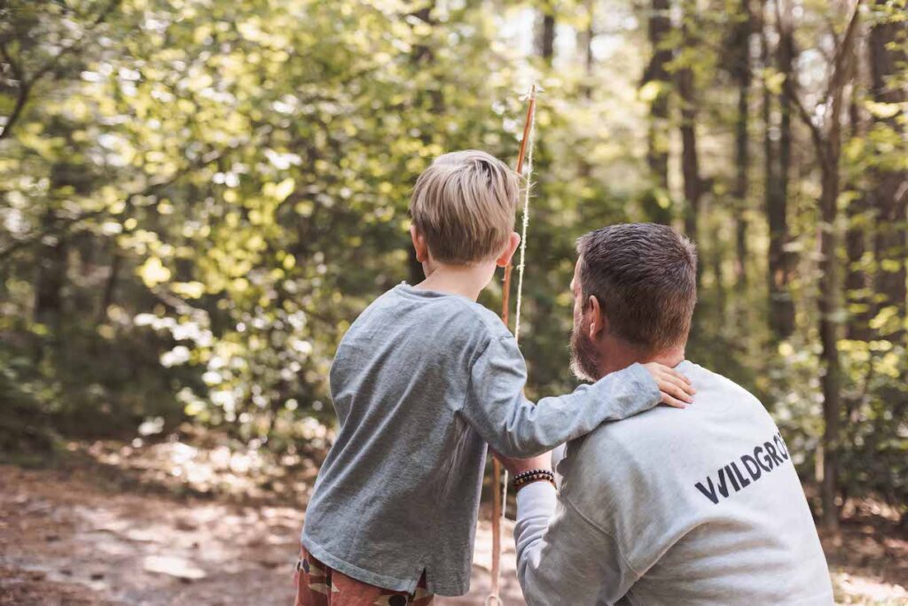 Een man met een "WILDGRAM"-sweatshirt knielt naast een kind dat een stok vasthoudt in het bos. Het kind, met kort blond haar, kijkt naar de bomen alsof het zijn volgende hutten bouwen-avontuur plant. Het zonlicht filtert zachtjes door de bladeren en werpt een speelse gloed.