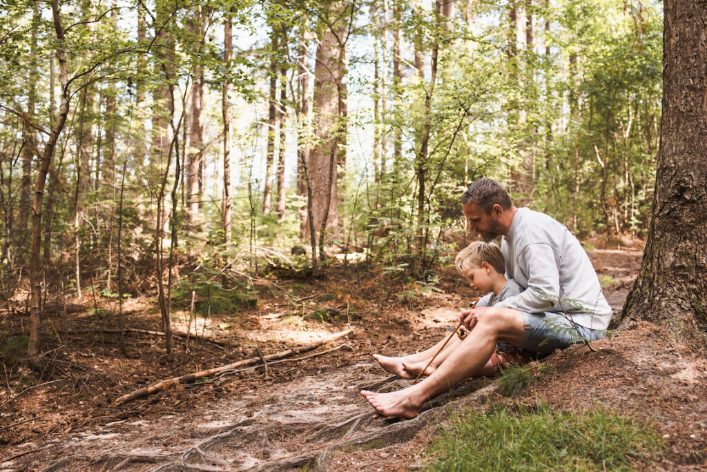 Een man en een kind zitten samen op een schaduwrijke bosbodem, omringd door hoge bomen en groen. Het kind leunt tegen de man aan, beiden op blote voeten, genietend van een vredig ouderlijk moment in de natuur tijdens hun hutten bouwen avontuur.