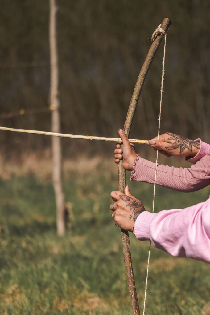 Een persoon met getatoeëerde handen en een roze mouw richt een handgemaakte houten boog naar buiten, waarbij hij de pees met de pijl erop naar achteren trekt tegen een met gras begroeide, wazige achtergrond.