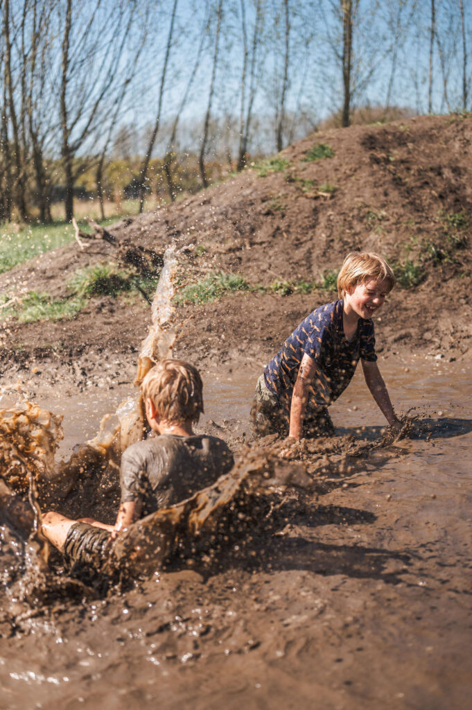 Twee jongens spelen vrolijk buiten in een modderplas, met opspattend water en modder onder een helderblauwe lucht en bomen op de achtergrond.